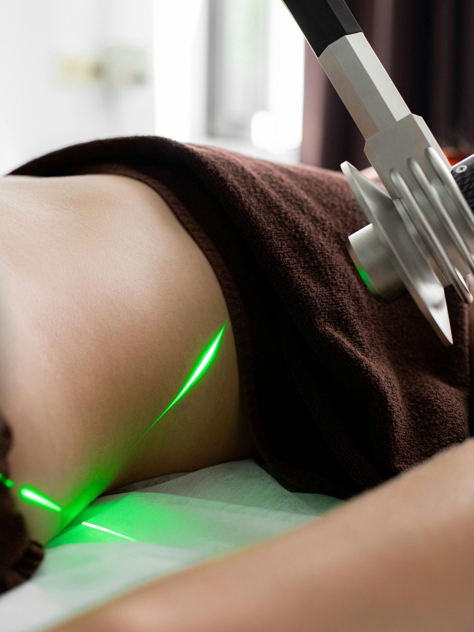 Crop anonymous patient with towel lying under green lasers of modern fat loss machine during procedure in light medical clinic